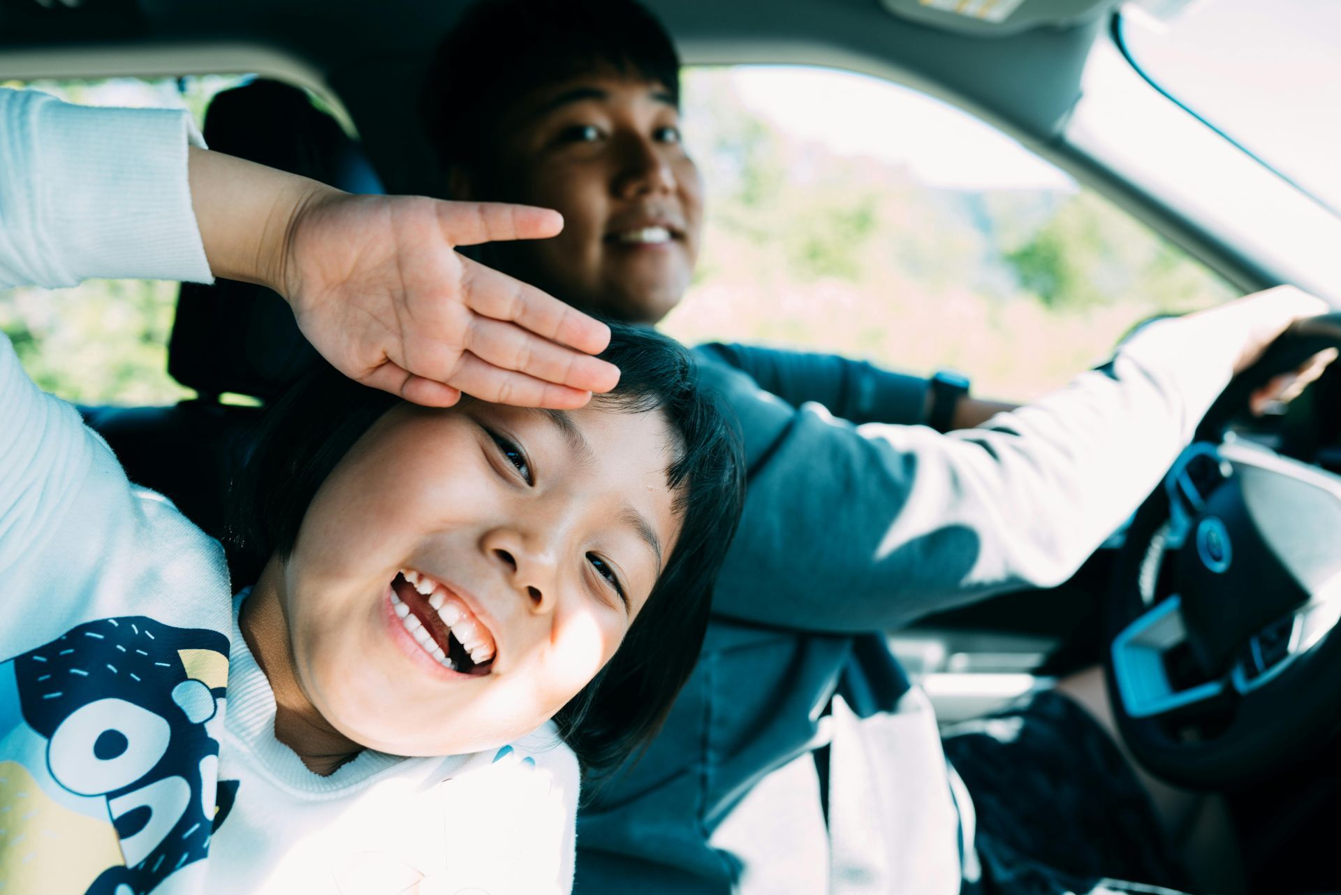 A child smiles while shading their eyes with one hand in the passenger seat of a car, with the driver visible in the back.
