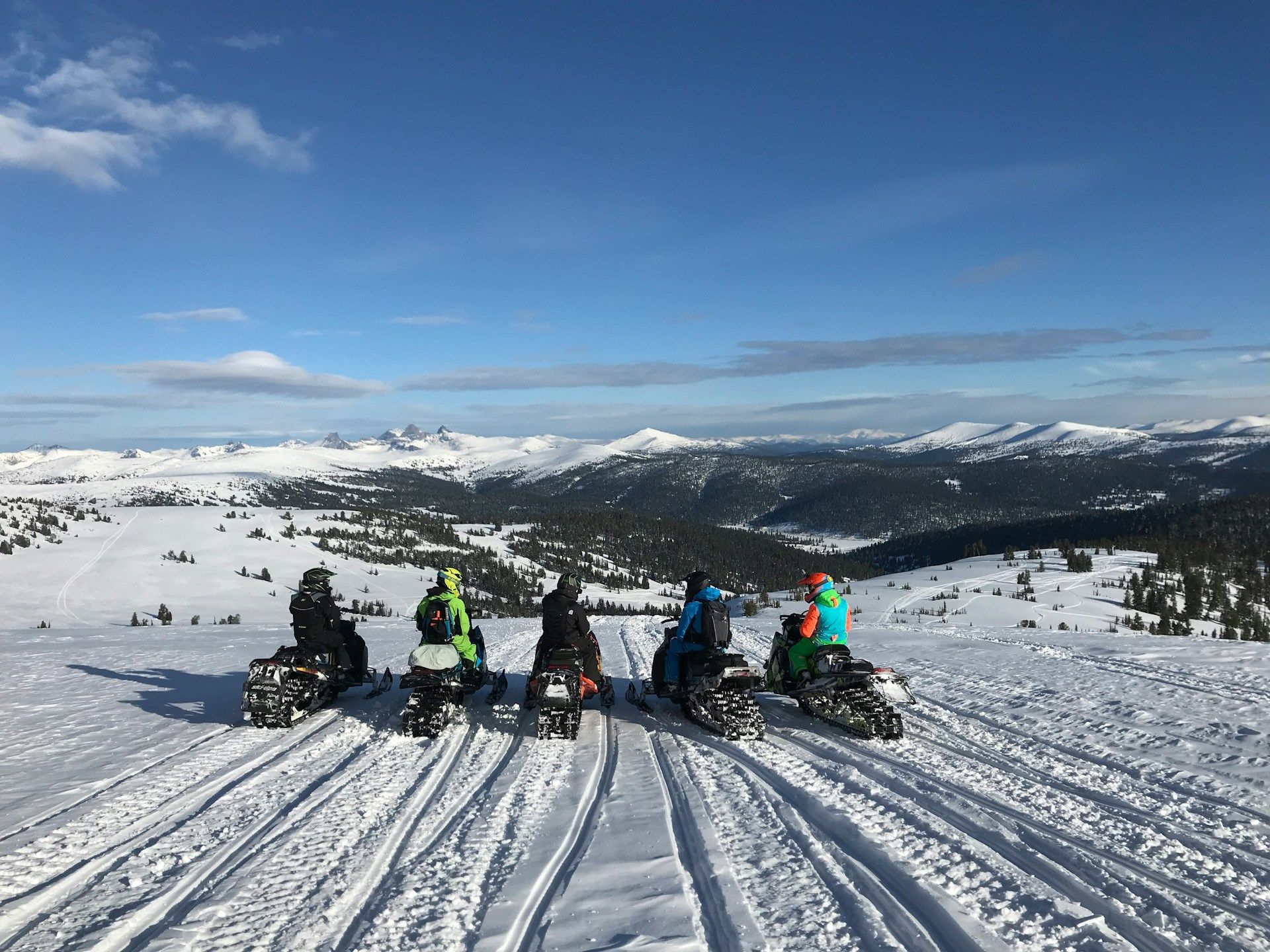 Five people on snowmobiles pause on a snowy ridge, facing a vast mountain landscape under a clear blue sky.