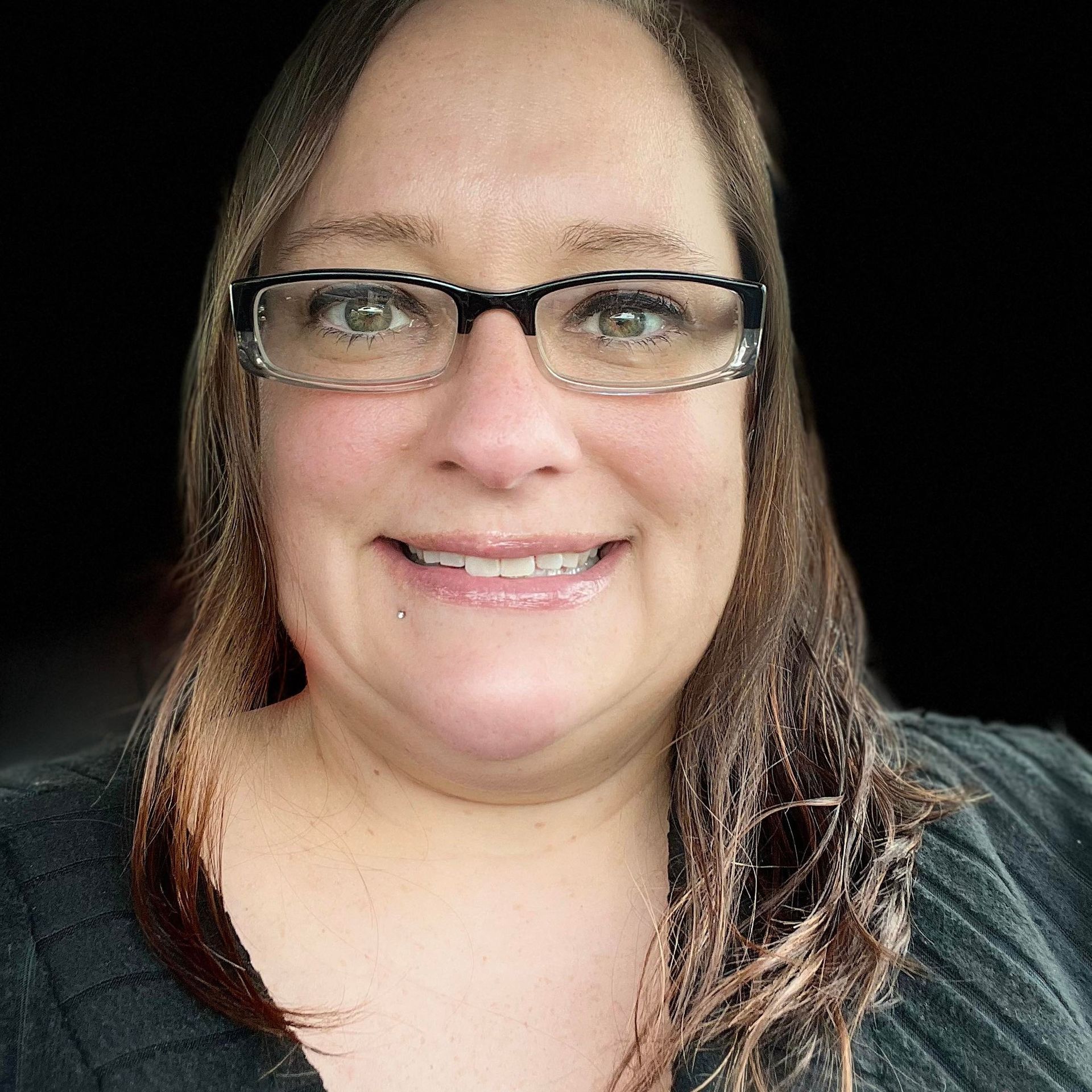 Woman with glasses smiles; brown hair, black shirt; against a dark background.