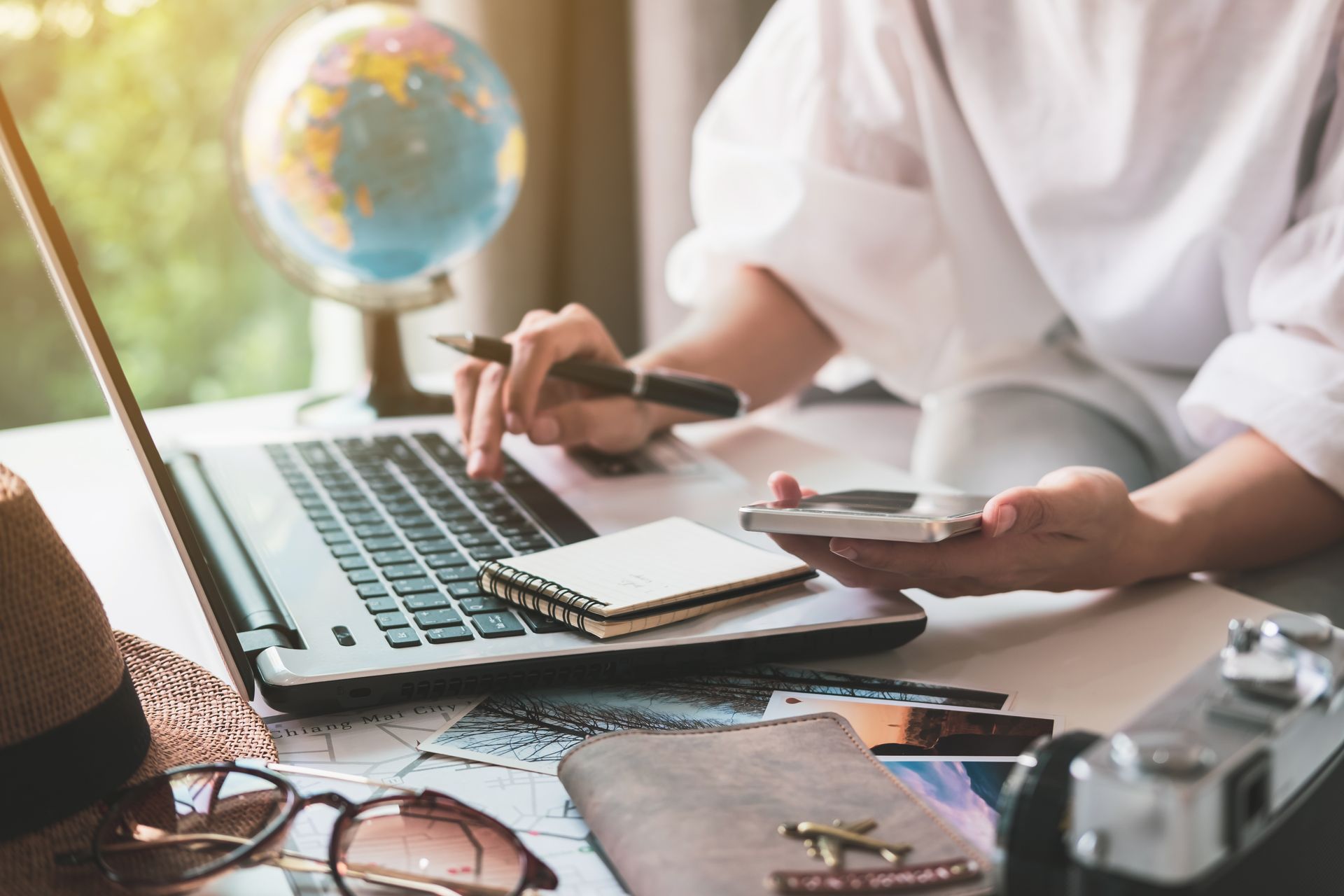 Person working on laptop at desk with travel items: globe, camera, sunglasses.