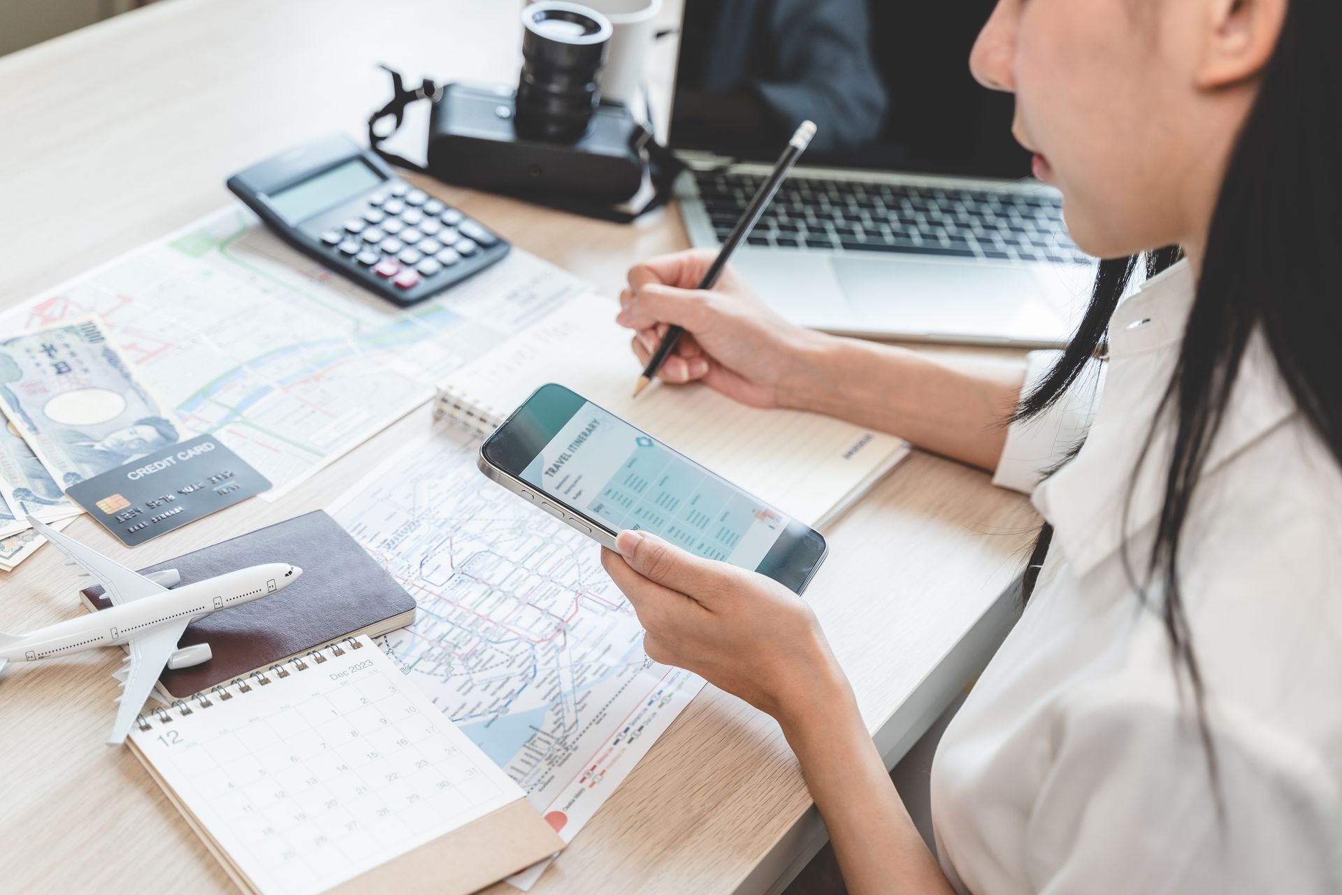 Woman planning a trip at desk with phone, notepad, map, calculator, camera, laptop, model airplane.