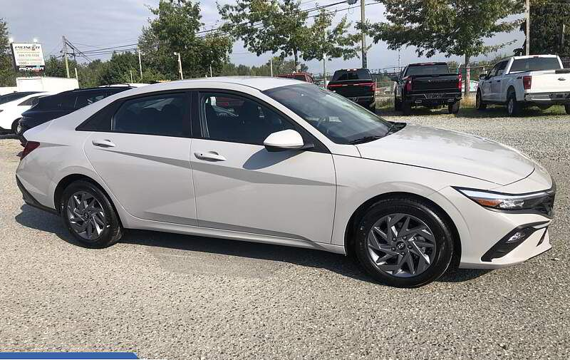 Light gray sedan parked on gravel, with other vehicles and trees visible in the background.
