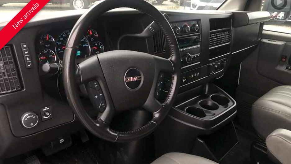 Interior of a GMC van, dashboard view. Black steering wheel with GMC logo, center console with cup holders.