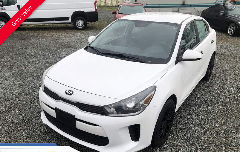 White Kia Rio parked on a gravel lot; other vehicles visible in the background.