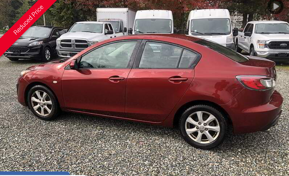 Red Mazda sedan parked outside a car dealership, with other vehicles in the background.