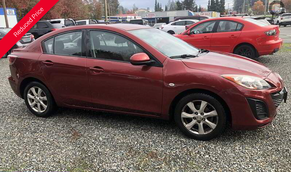 Red Mazda 3 sedan parked on gravel lot. Another red car is in the background.