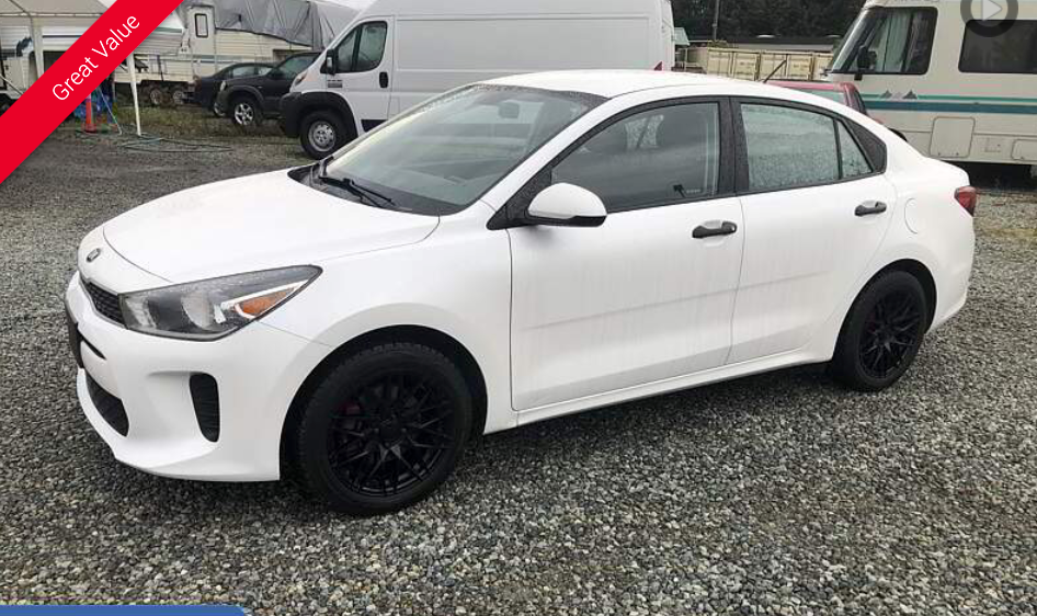 White Kia Rio sedan with black wheels parked on gravel.