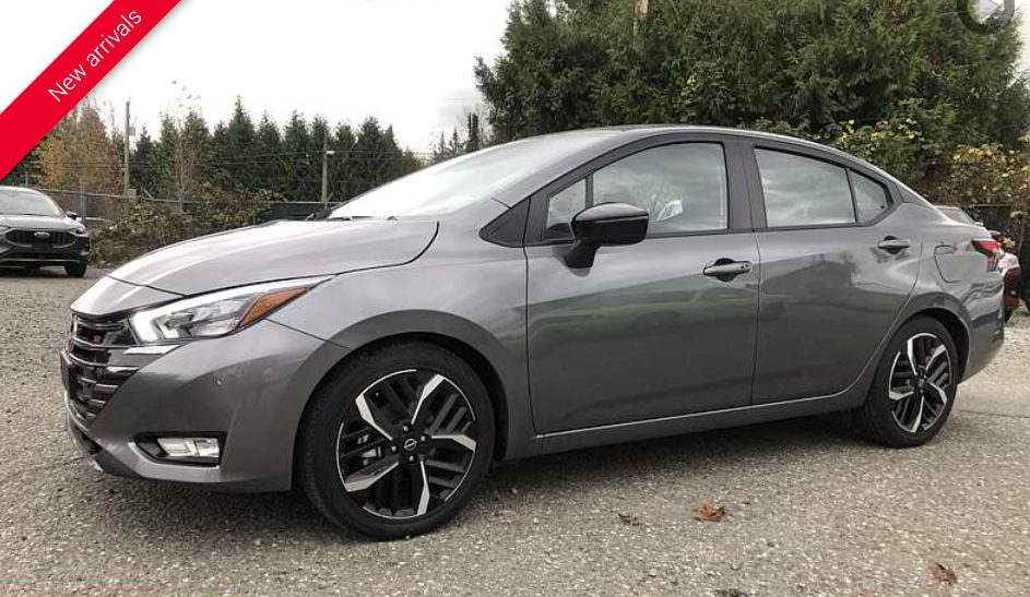 Gray Nissan Versa sedan parked on gravel in front of trees.