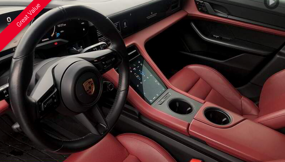 Black steering wheel and red leather interior of a Porsche Taycan. Center console has cup holders and a touchscreen.