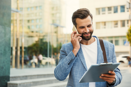 Man in blue blazer talks on phone, smiles while holding a clipboard outside a modern building.
