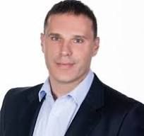 Man wearing a blue shirt and blazer, smiling, against a white background.