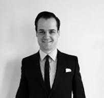 Man in black suit smiles. White wall background.