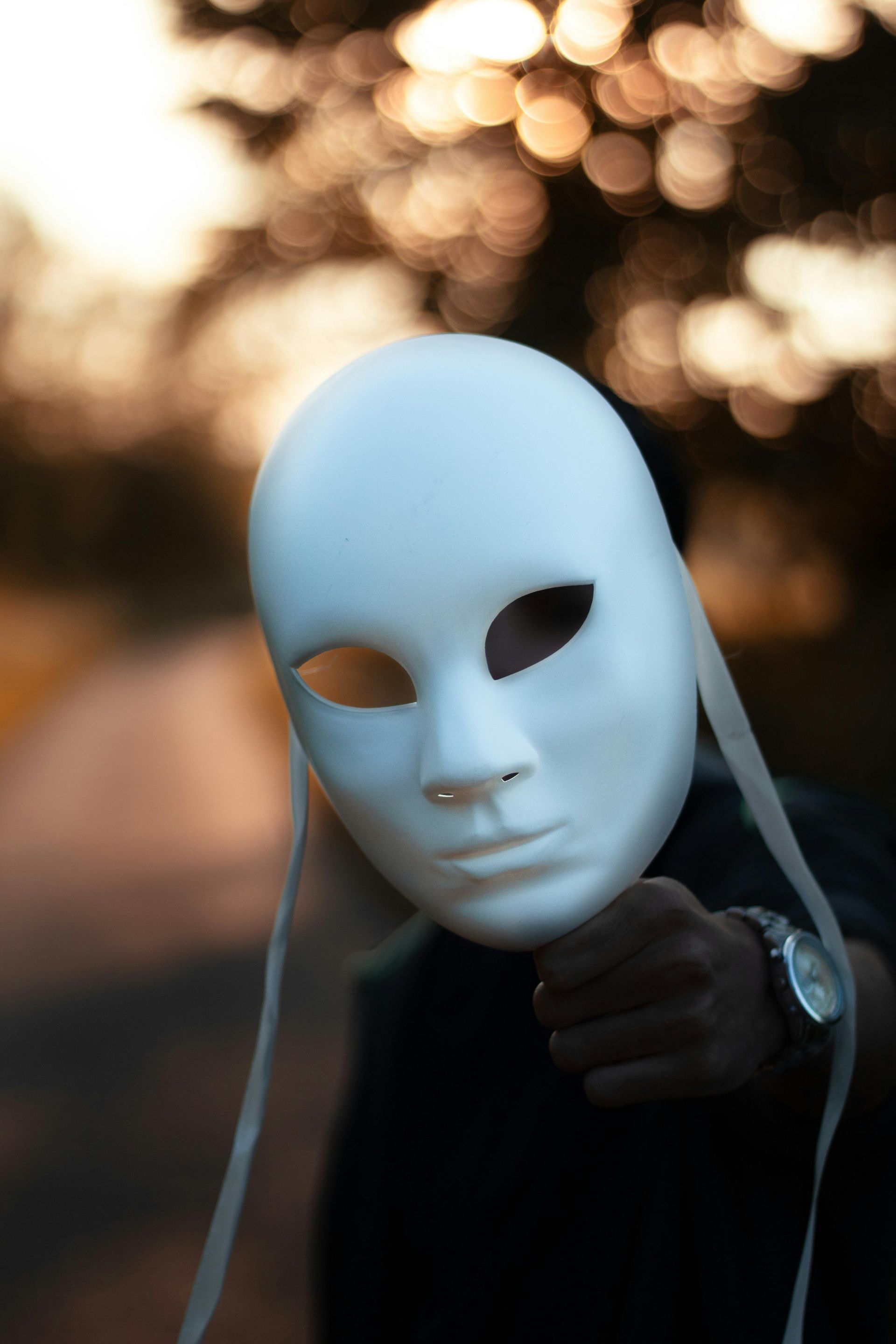 Person holding up a white mask with neutral expression, blurry background of trees and bokeh.
