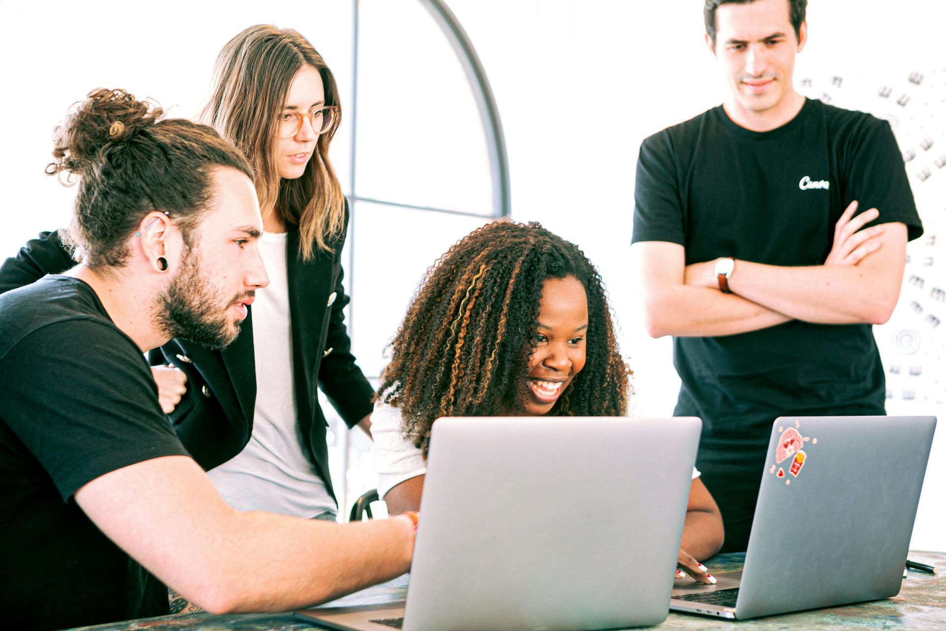 Four people collaborating around two laptops; smiling woman points, others observe.