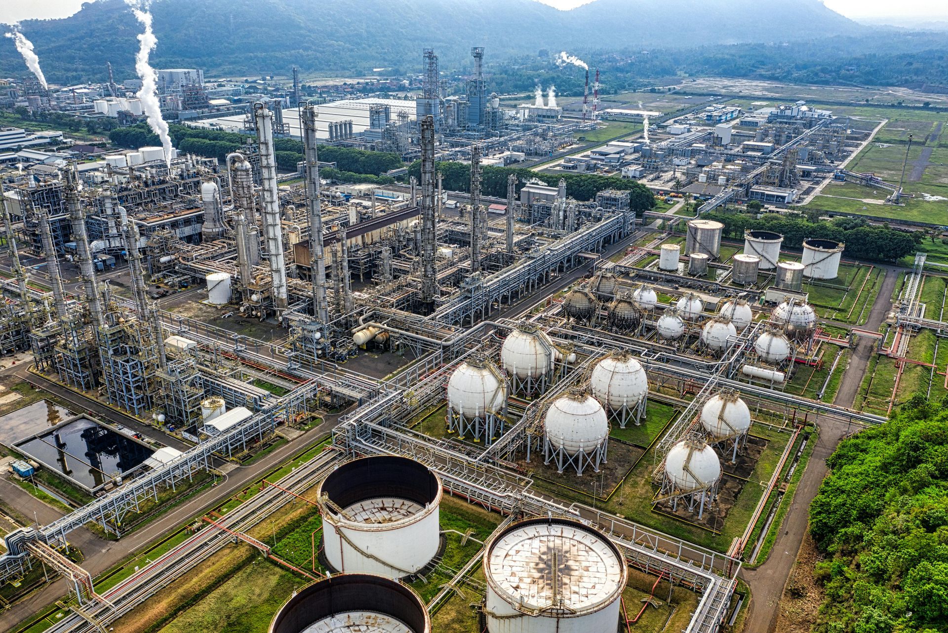 Aerial view of a large oil refinery complex with storage tanks, pipes, and smokestacks against a green landscape.