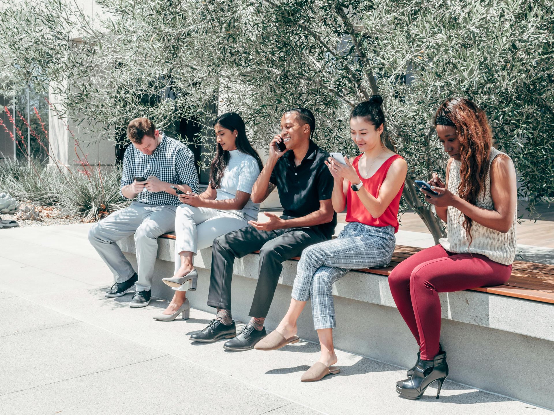 Five people sit on a bench outdoors, using phones. One is on a call, another claps. Sunny day.