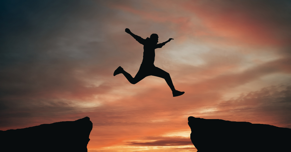 Silhouette of a person leaping between two cliffs at sunset with an orange sky