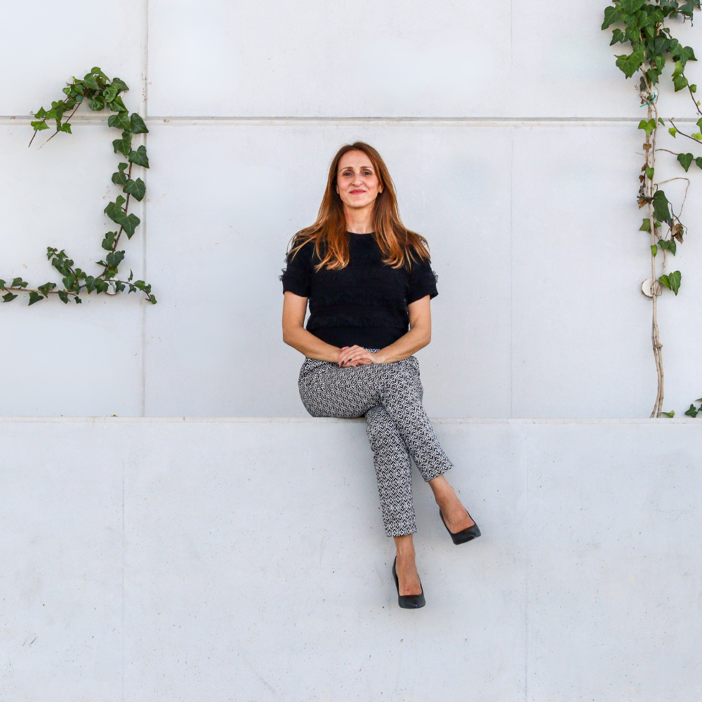 Woman sitting on a white wall, wearing black top and patterned pants, with ivy.