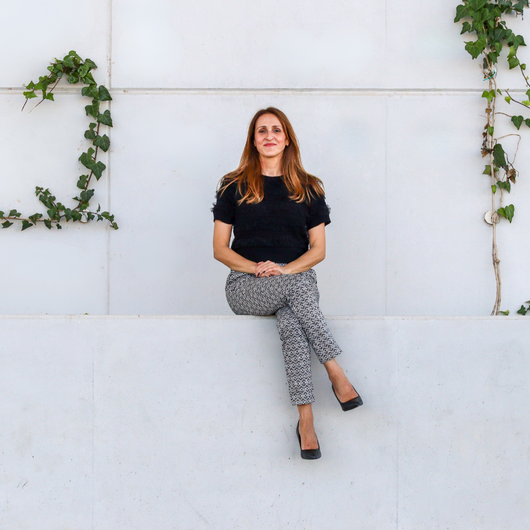 Woman sitting on a white wall, wearing black top and patterned pants, with ivy.