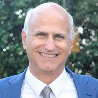 Man with light skin smiling, wearing a blue blazer, white shirt, and gray tie.