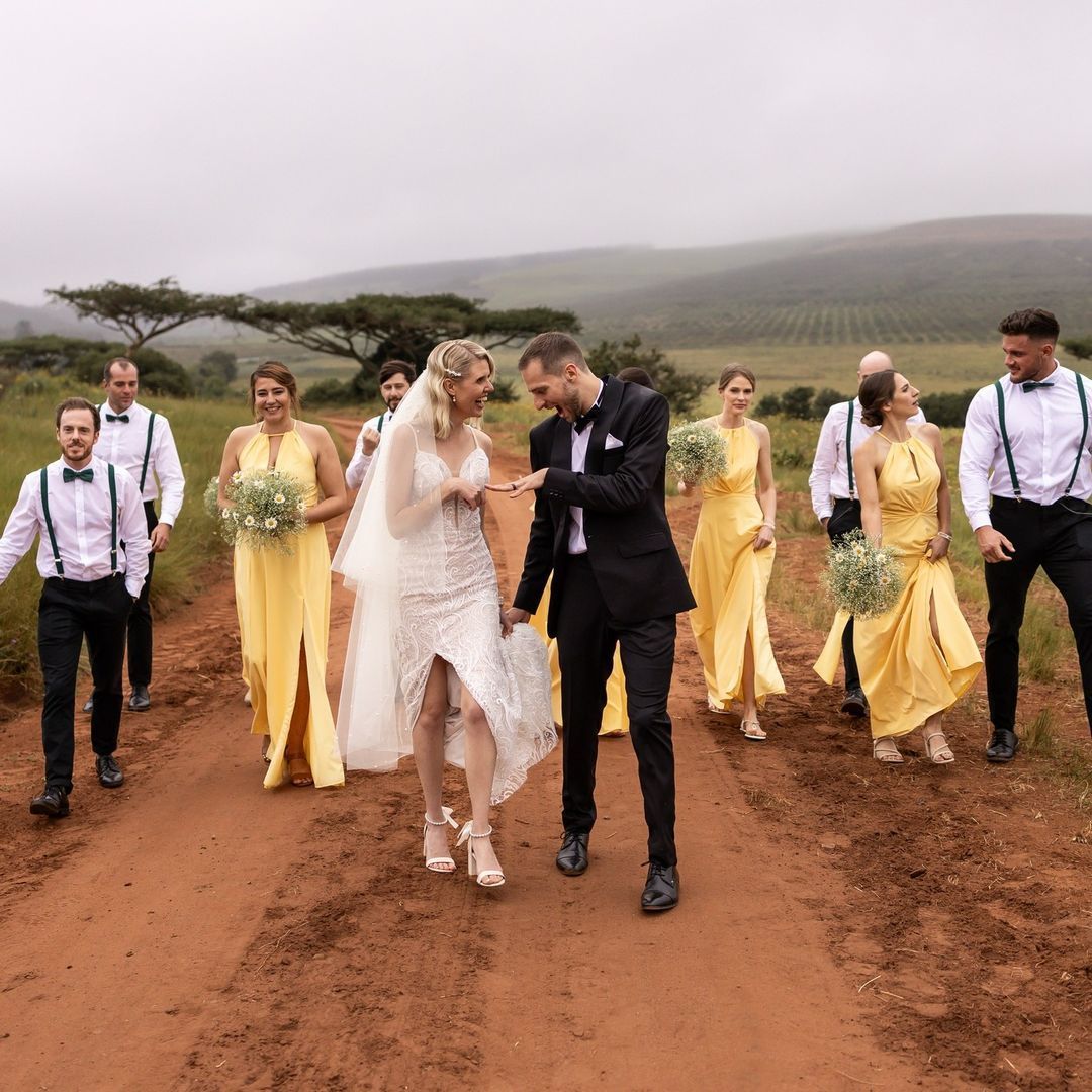A bride and groom are walking down a dirt road with their wedding party.