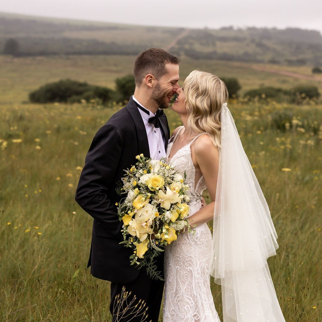 A bride and groom are kissing in a field while the bride is holding a bouquet of flowers.