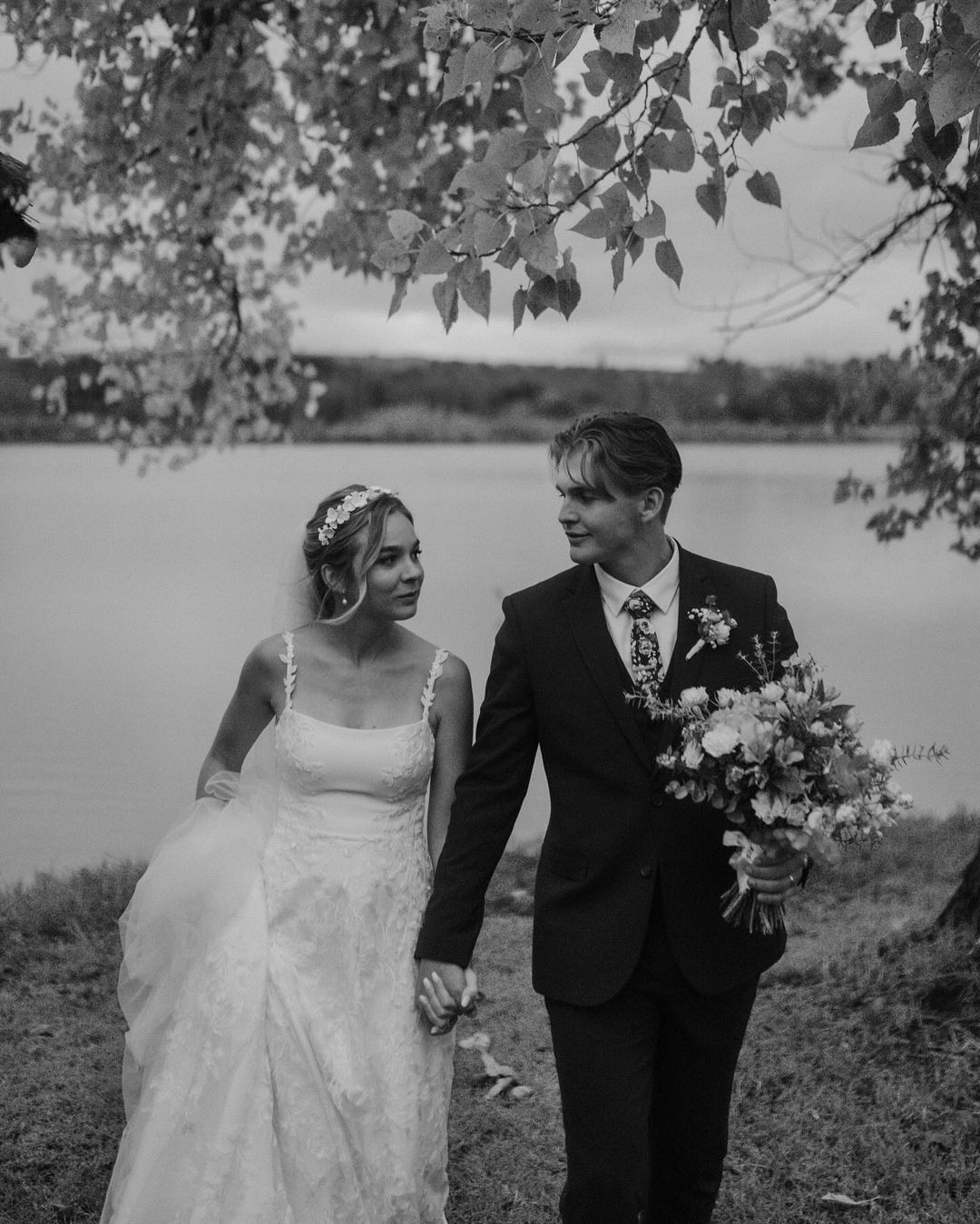 A bride and groom are walking by a lake holding hands.