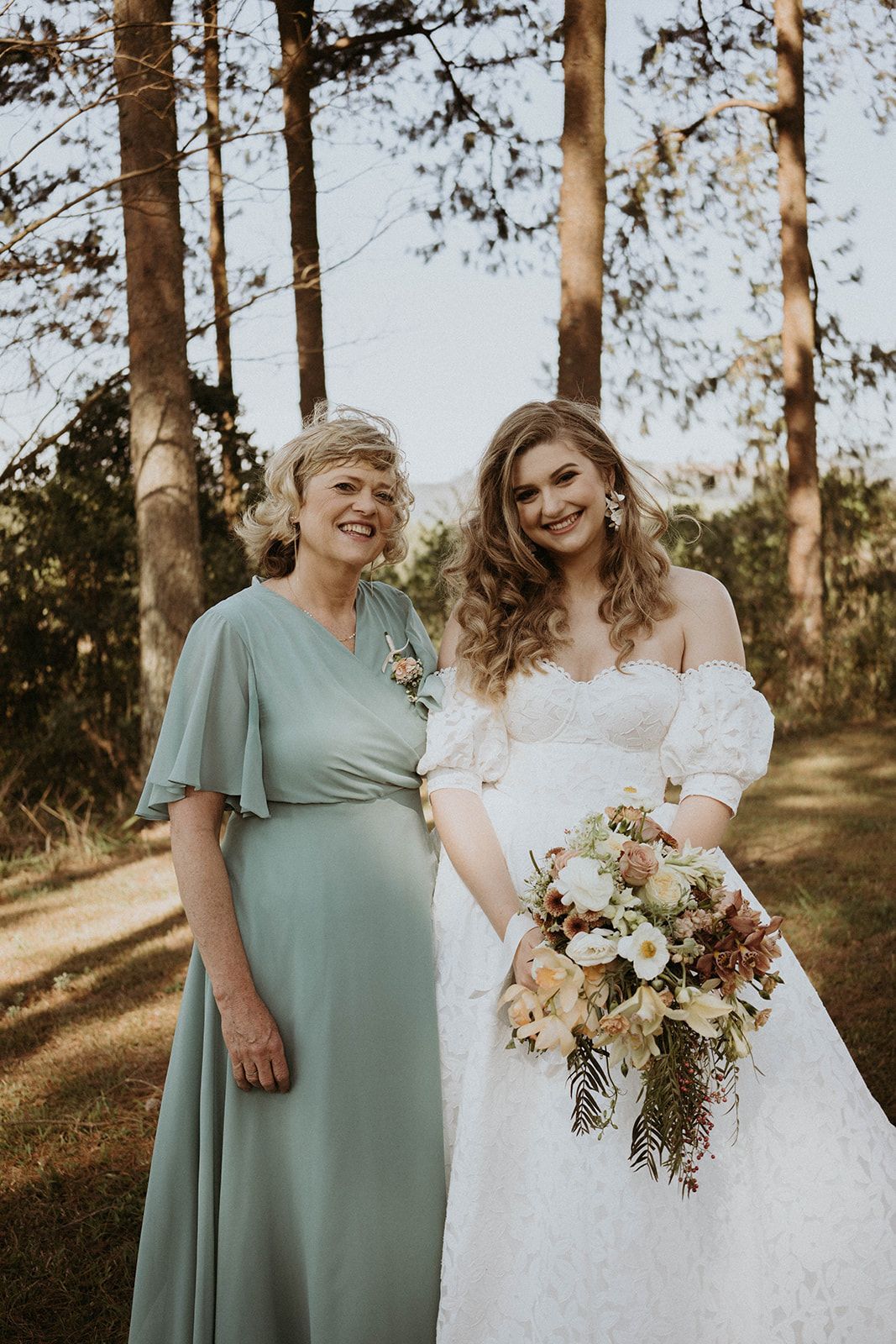 A bride and her mother are posing for a picture in the woods.