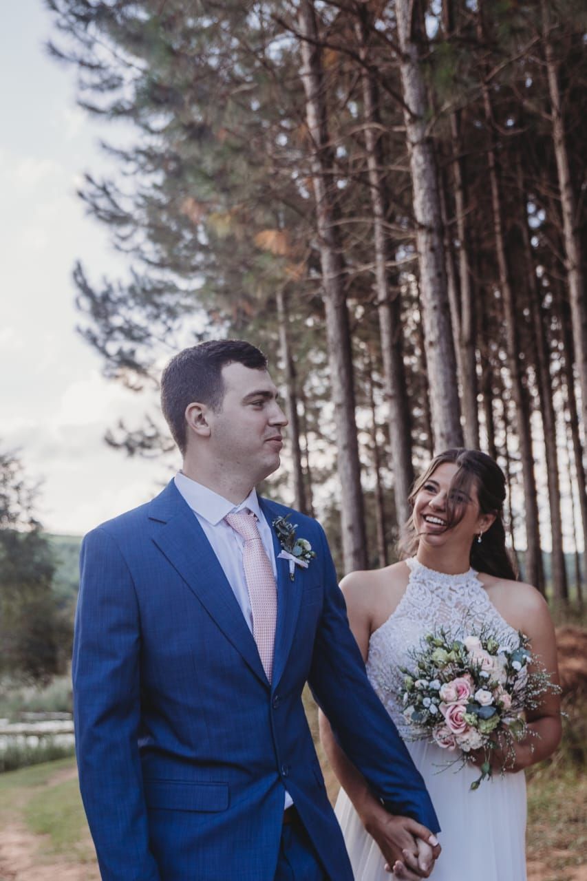 A bride and groom are walking through a forest holding hands.
