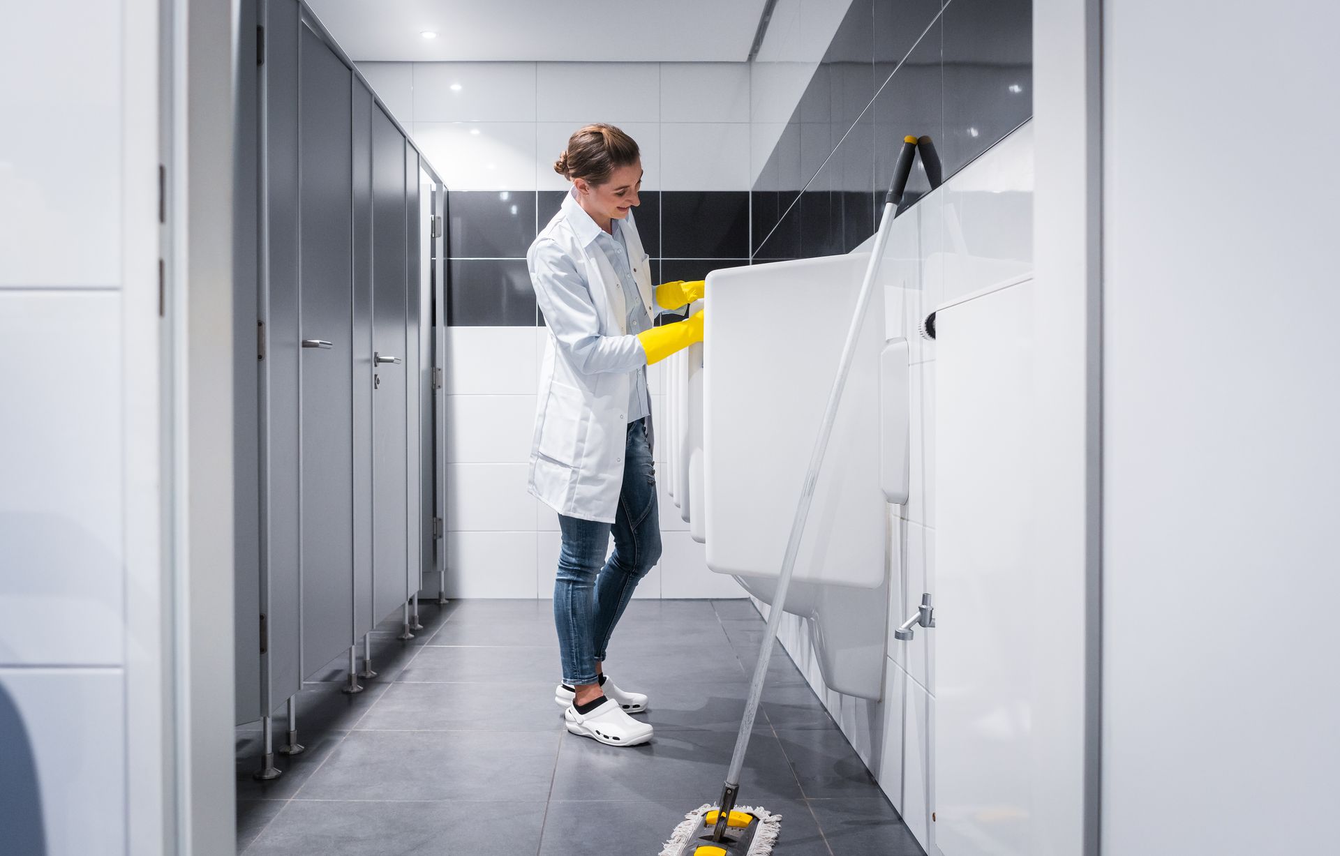 Woman in lab coat, yellow gloves mops a public restroom with white stalls and walls.