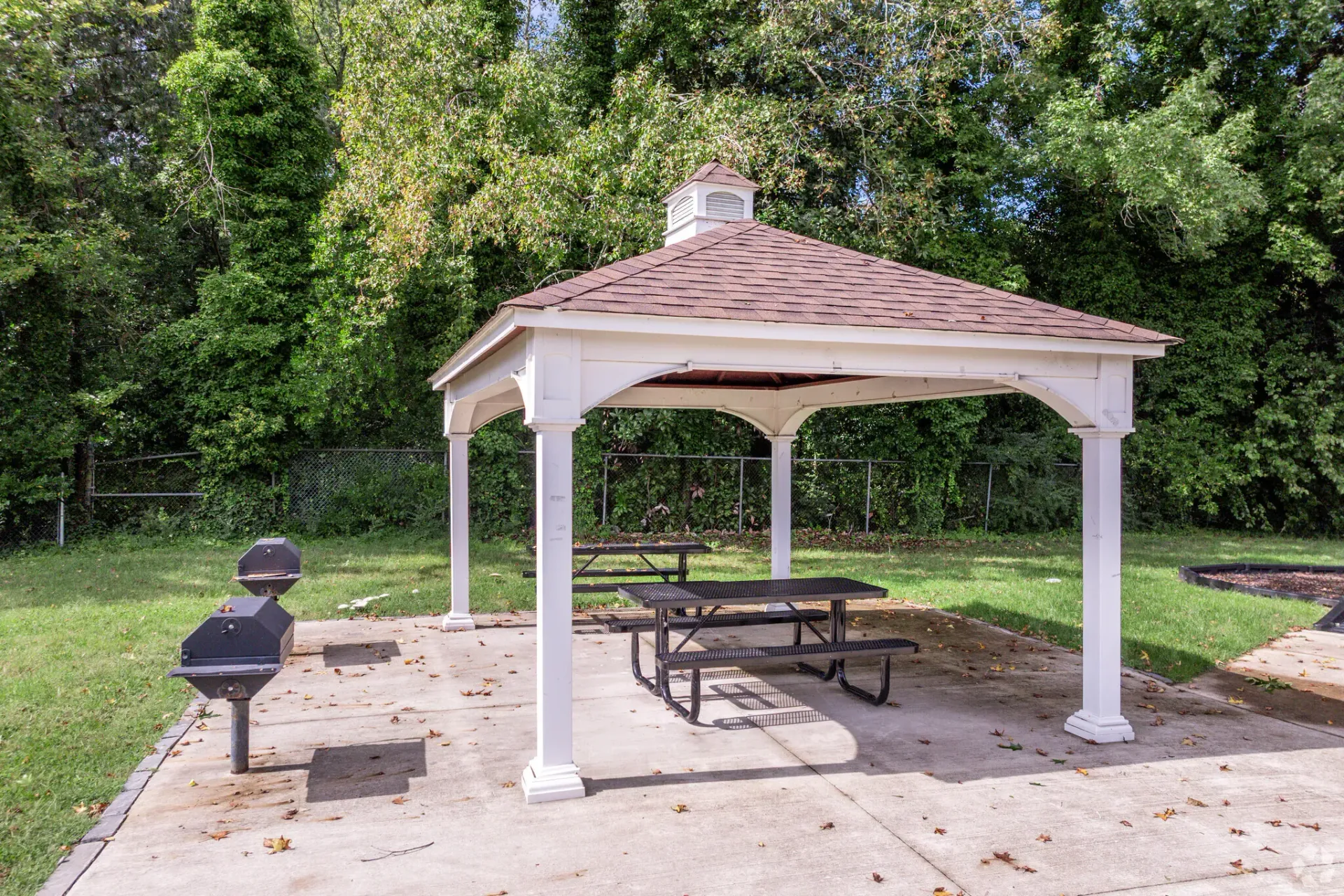 White gazebo with red roof, picnic tables, and grills in a grassy, tree-filled community area. Enjoy a variety of onsite community amenities at Harborstone Apartments in Newport News, VA. 