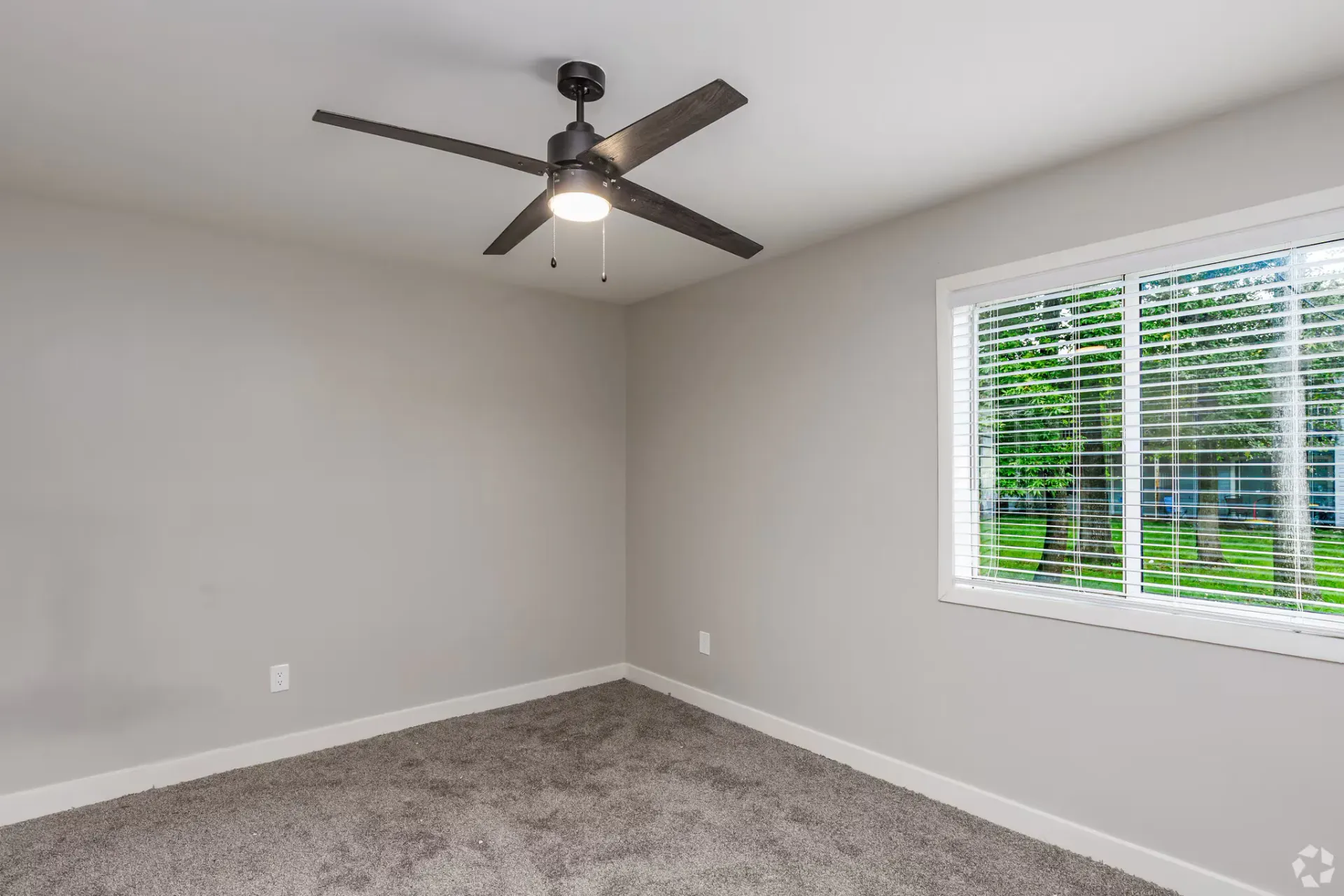 Empty bedroom with gray walls, carpet, ceiling fan, and a window with blinds. Contact our leasing team at Harborstone Apartments today to schedule a tour of our studio, one-, and two-bedroom apartments in Newport News, VA. 