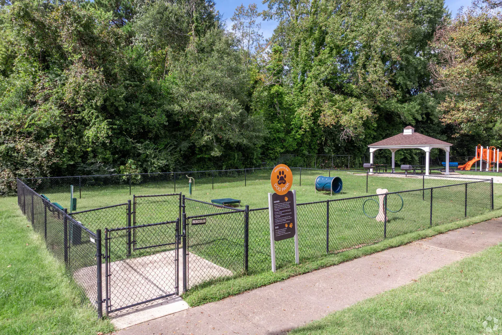 Fenced dog park with a sign, gazebo, and playground beyond, bordered by trees. Enjoy a pet-friendly community at Harborstone Apartments in Newport News, VA. 