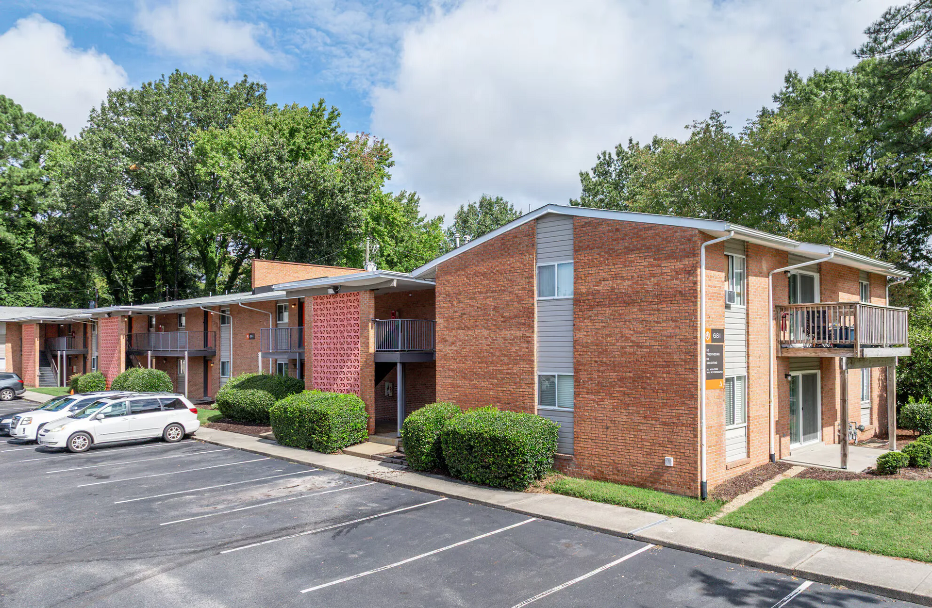 Exterior view of a brick apartment building with a parking lot and trees. Welcome home to Harborstone Apartments, offering studio, 1, and 2-bedroom apartments for rent in New Port News, VA.