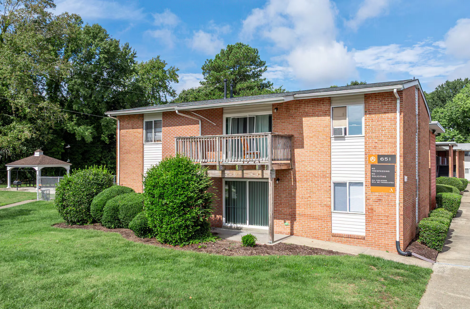 Two-story brick apartment building exterior with a wooden balcony, trimmed shrubs, and green lawn. Welcome home to Harborstone Apartments, offering studio, 1, and 2-bedroom apartments for rent in New Port News, VA.