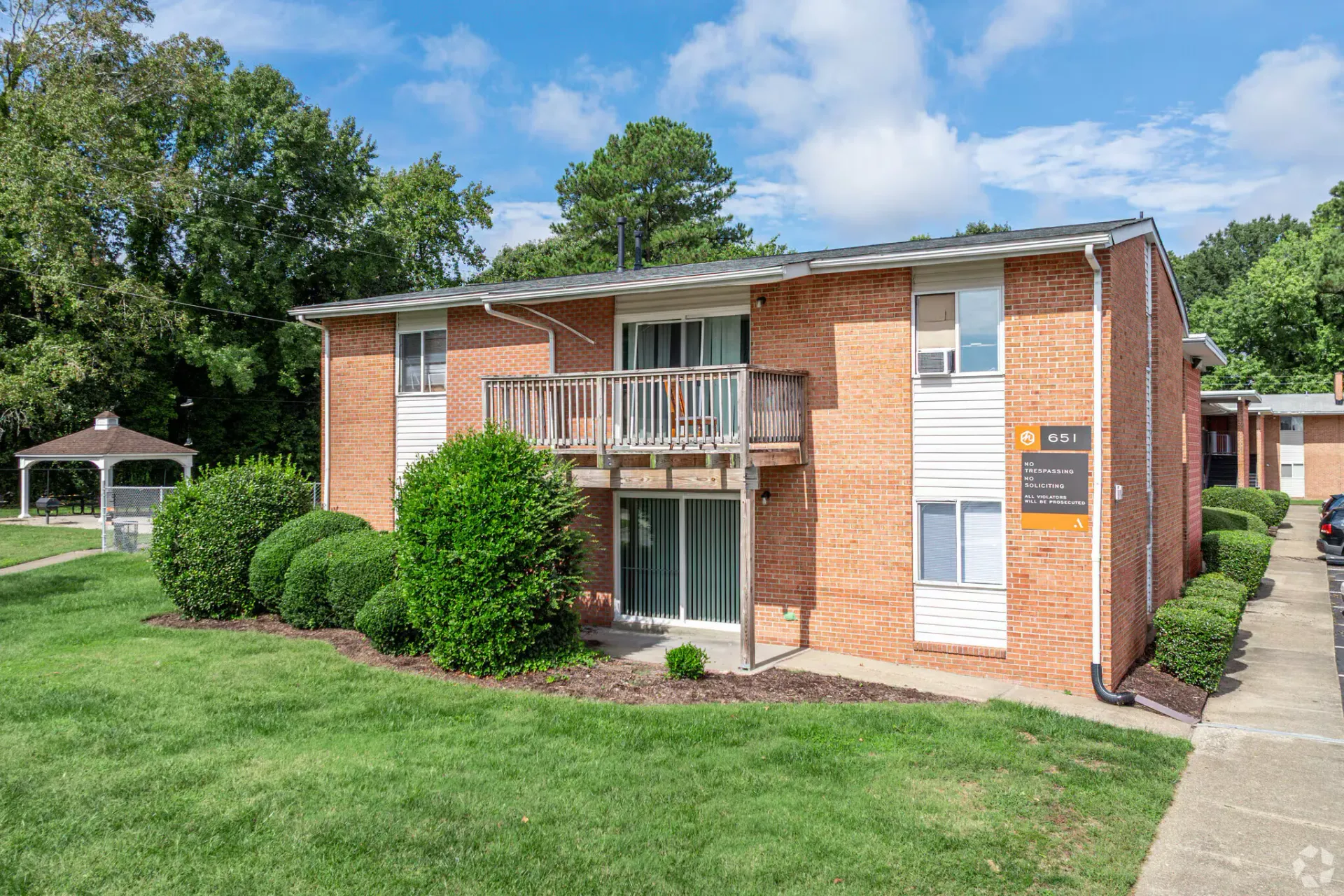 Two-story brick apartment building exterior with a wooden balcony, trimmed shrubs, and green lawn. Welcome home to Harborstone Apartments, offering studio, 1, and 2-bedroom apartments for rent in New Port News, VA.