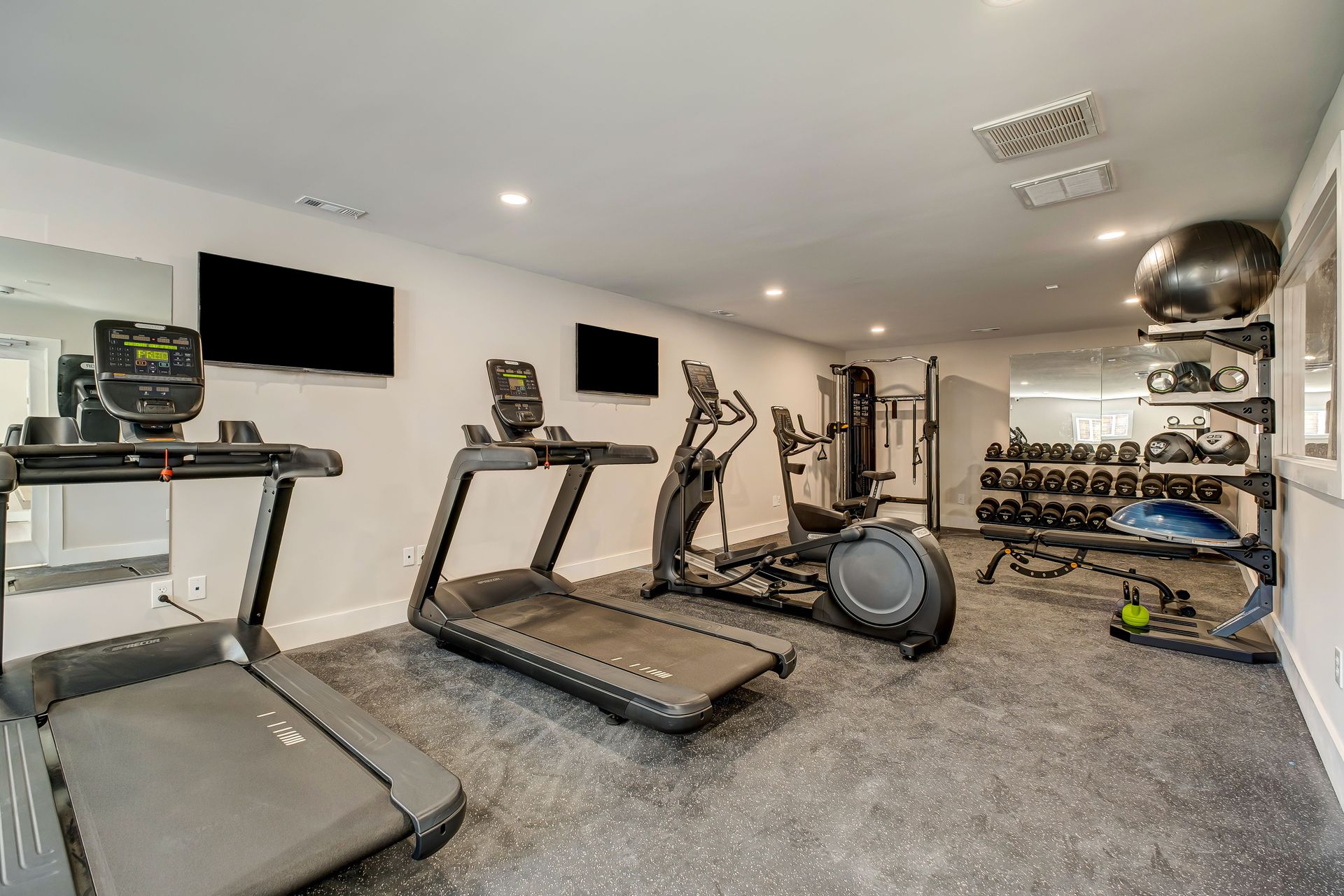 A fitness room featuring two treadmills, an elliptical, a stationary bike, and a rack of dumbbells on a gray carpet.