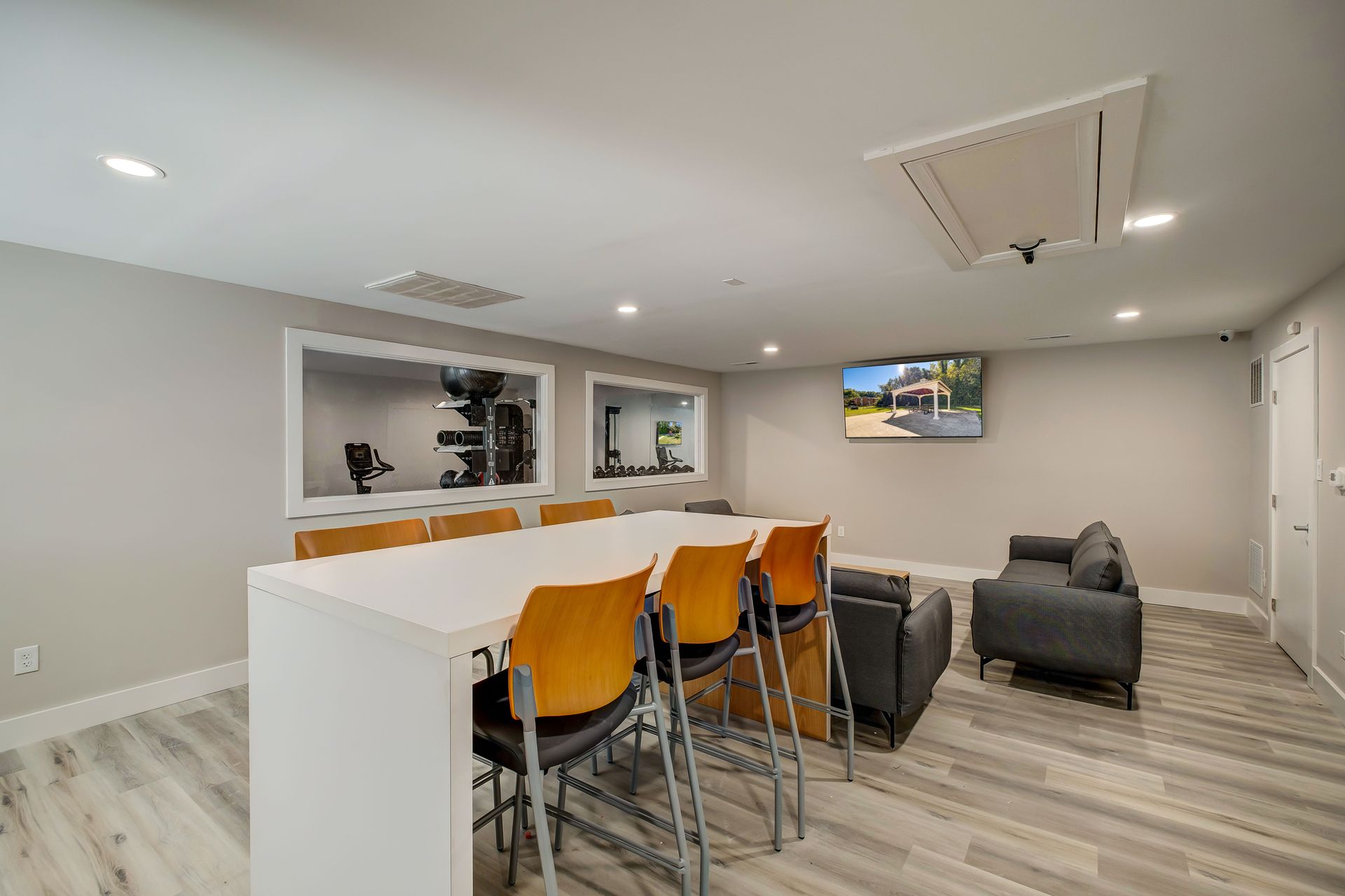 A modern lounge with a long white table, yellow chairs, a grey sofa, and a wall-mounted TV in a light-colored room.