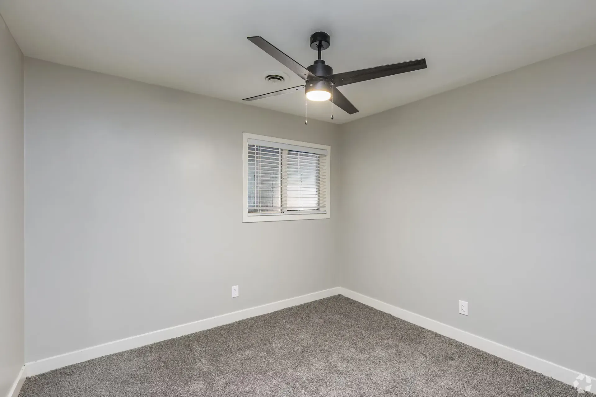Empty bedroom with light gray walls, white trim, window blinds, gray carpet, and a ceiling fan. Contact our leasing team at Harborstone Apartments today to schedule a tour of our studio, one-, and two-bedroom apartments in Newport News, VA. 