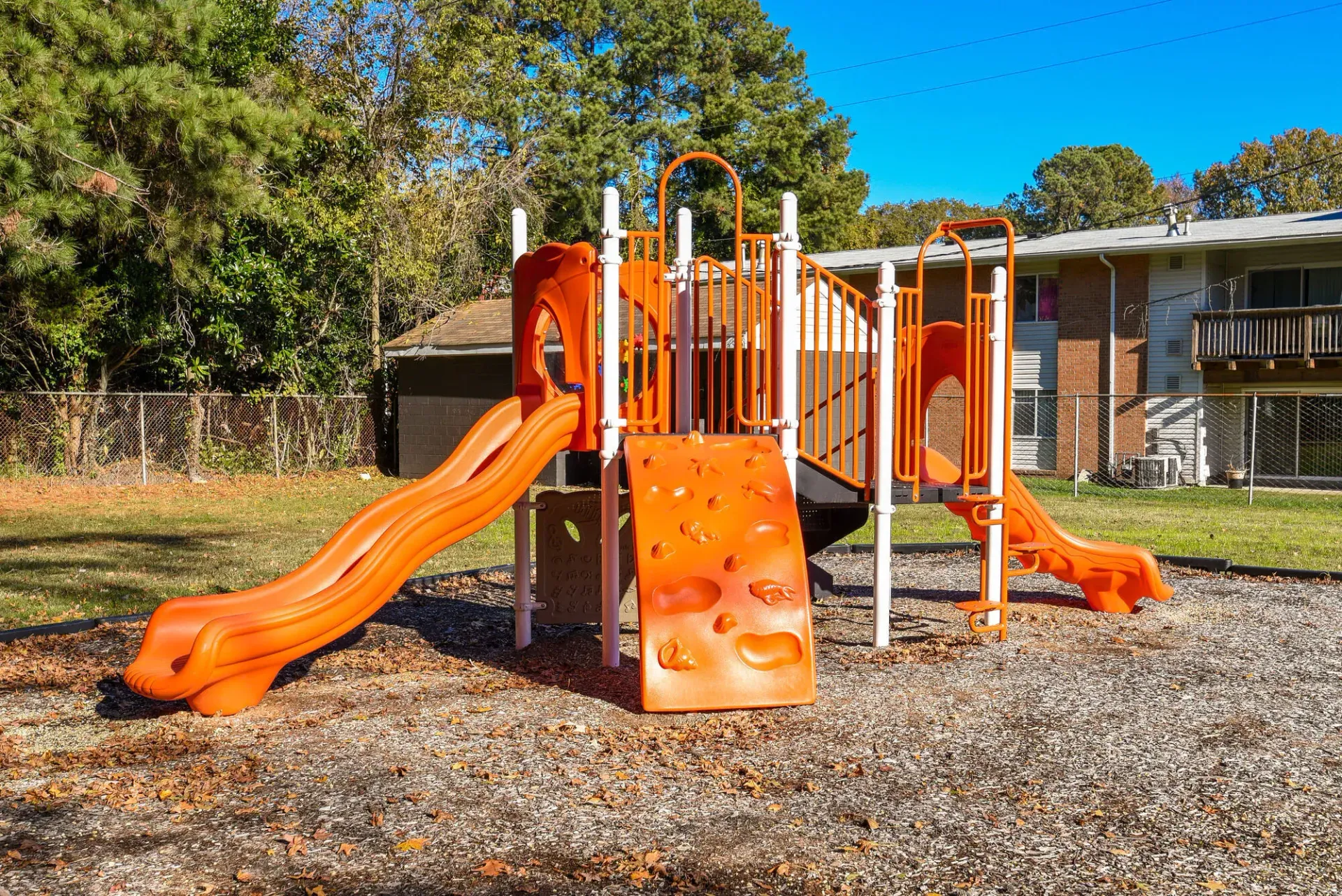 Orange playground structure with slides and climbing wall in a fenced apartment complex yard. Enjoy a variety of onsite community amenities at Harborstone Apartments in Newport News, VA. 