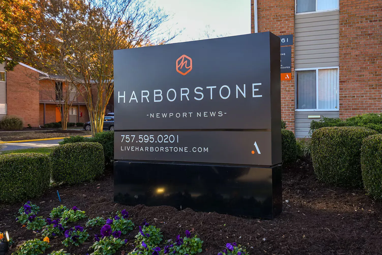 Large dark Harborstone sign in front of a brick apartment building with trimmed hedges and flowers. Welcome home to Harborstone Apartments, offering studio, 1, and 2-bedroom apartments for rent in New Port News, VA.