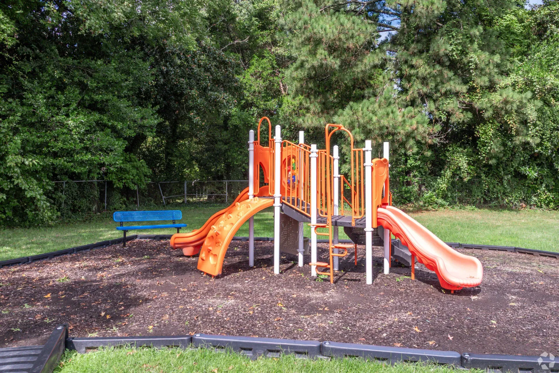 Bright orange playground structure with slides in a fenced, tree-filled area. Enjoy a variety of onsite community amenities at Harborstone Apartments in Newport News, VA. 
