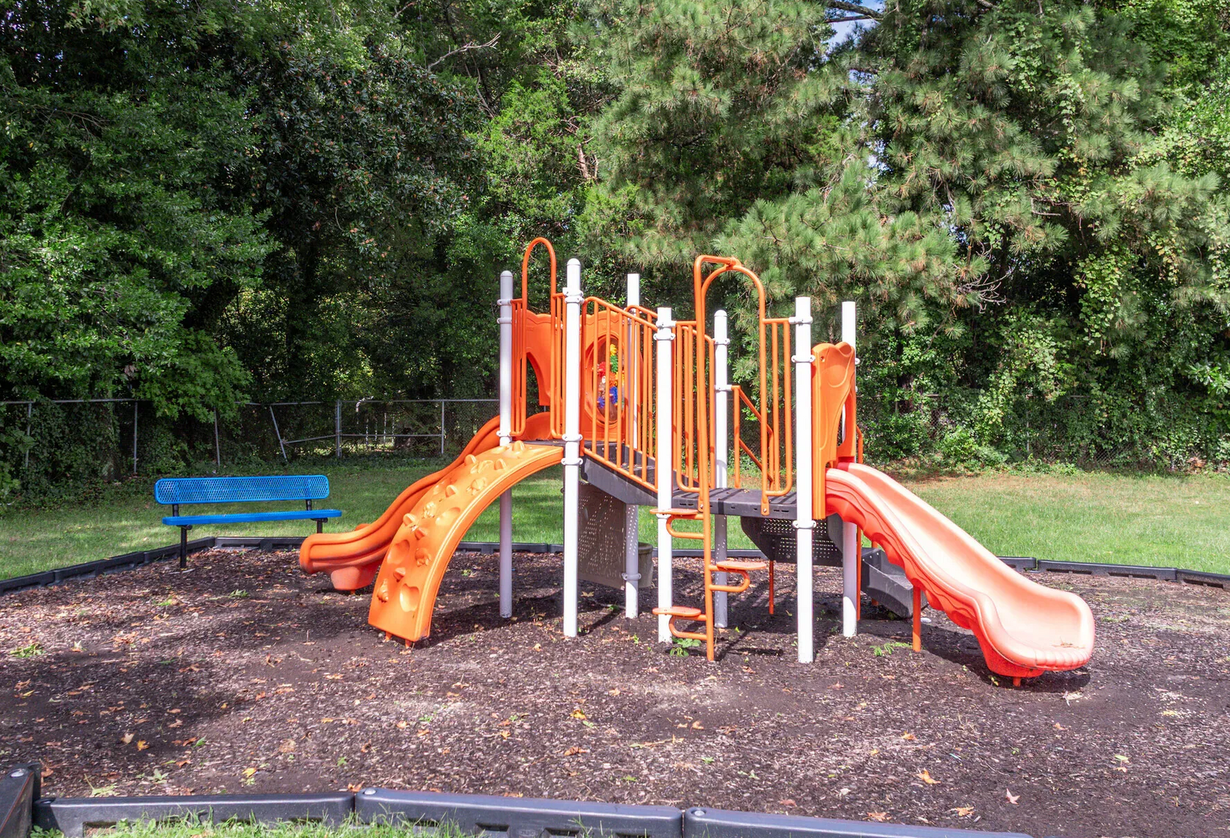 Bright orange playground structure with slides in a fenced, tree-filled area. Enjoy a variety of onsite community amenities at Harborstone Apartments in Newport News, VA. 