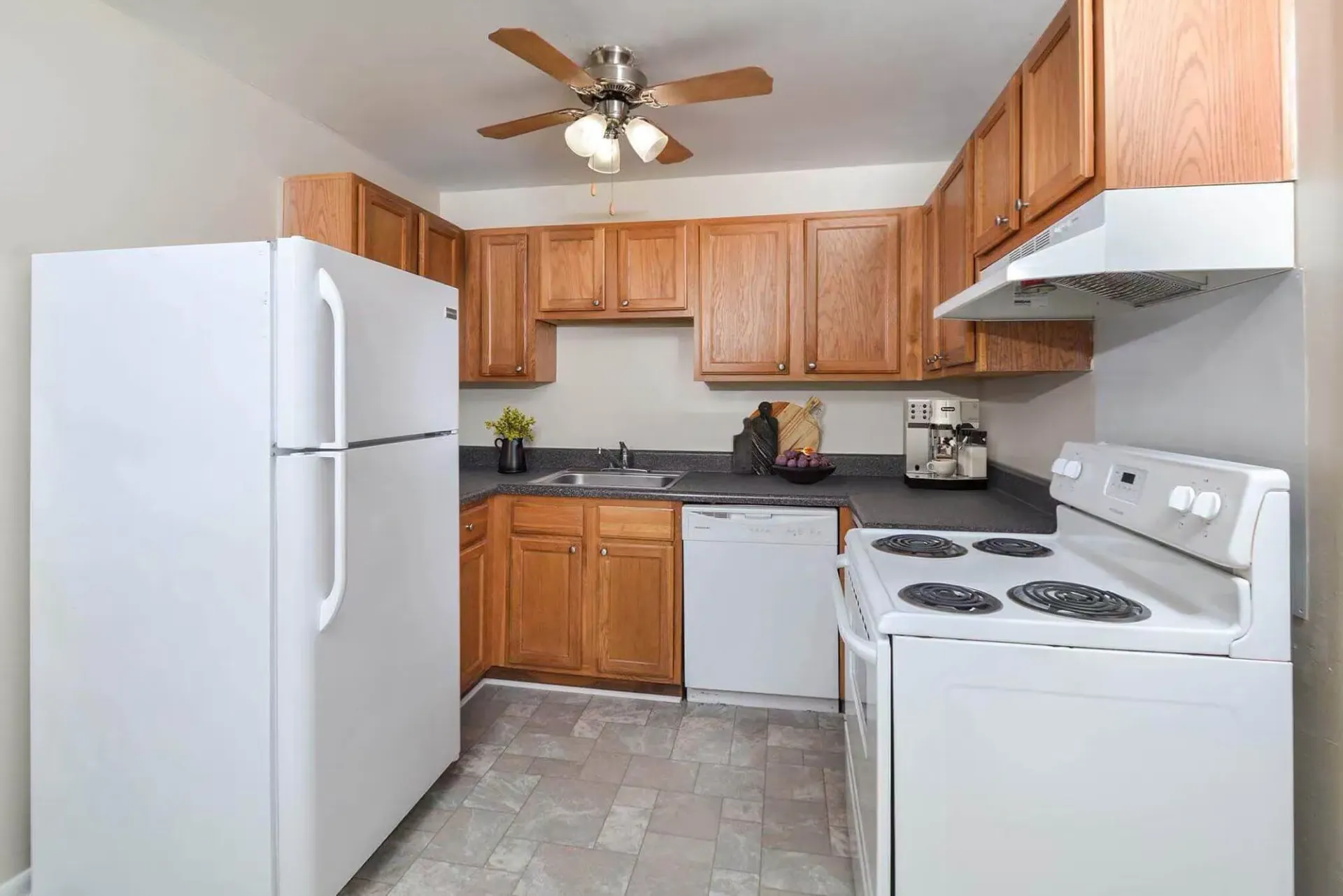 Compact apartment kitchen with wooden cabinets, white appliances, and a ceiling fan. Contact our leasing team at Harborstone Apartments today to schedule a tour of our studio, one-, and two-bedroom apartments in Newport News, VA. 