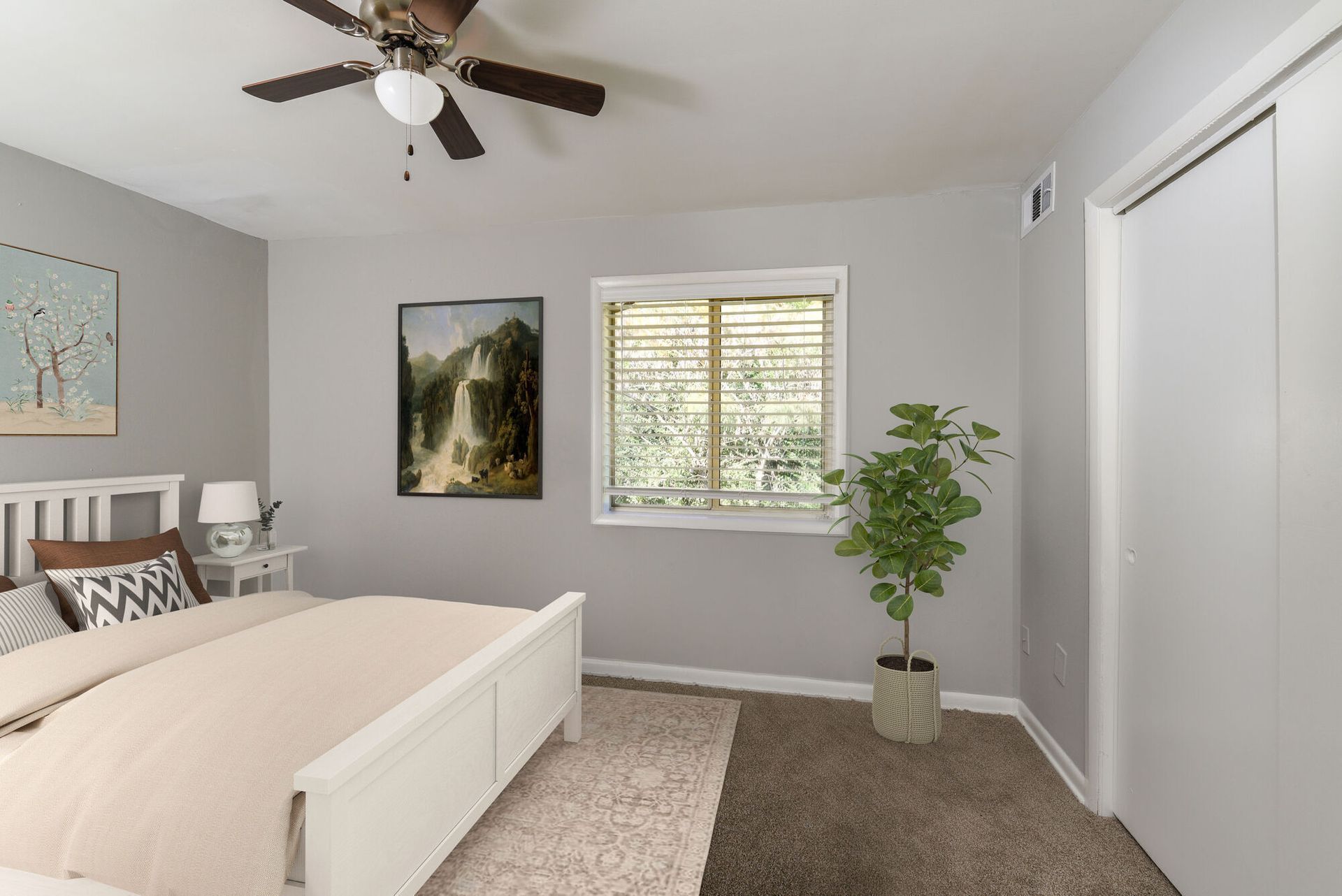 Bedroom scene with white bed frame, nightstand, ceiling fan, window, and potted plant. Contact our leasing team at Harborstone Apartments today to schedule a tour of our studio, one-, and two-bedroom apartments in Newport News, VA. 