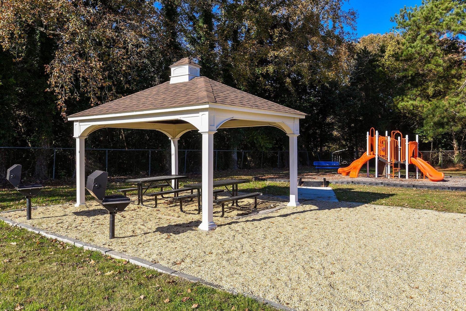 White pavilion with picnic tables and grills beside an orange playground set. Enjoy a variety of onsite community amenities at Harborstone Apartments in Newport News, VA. 