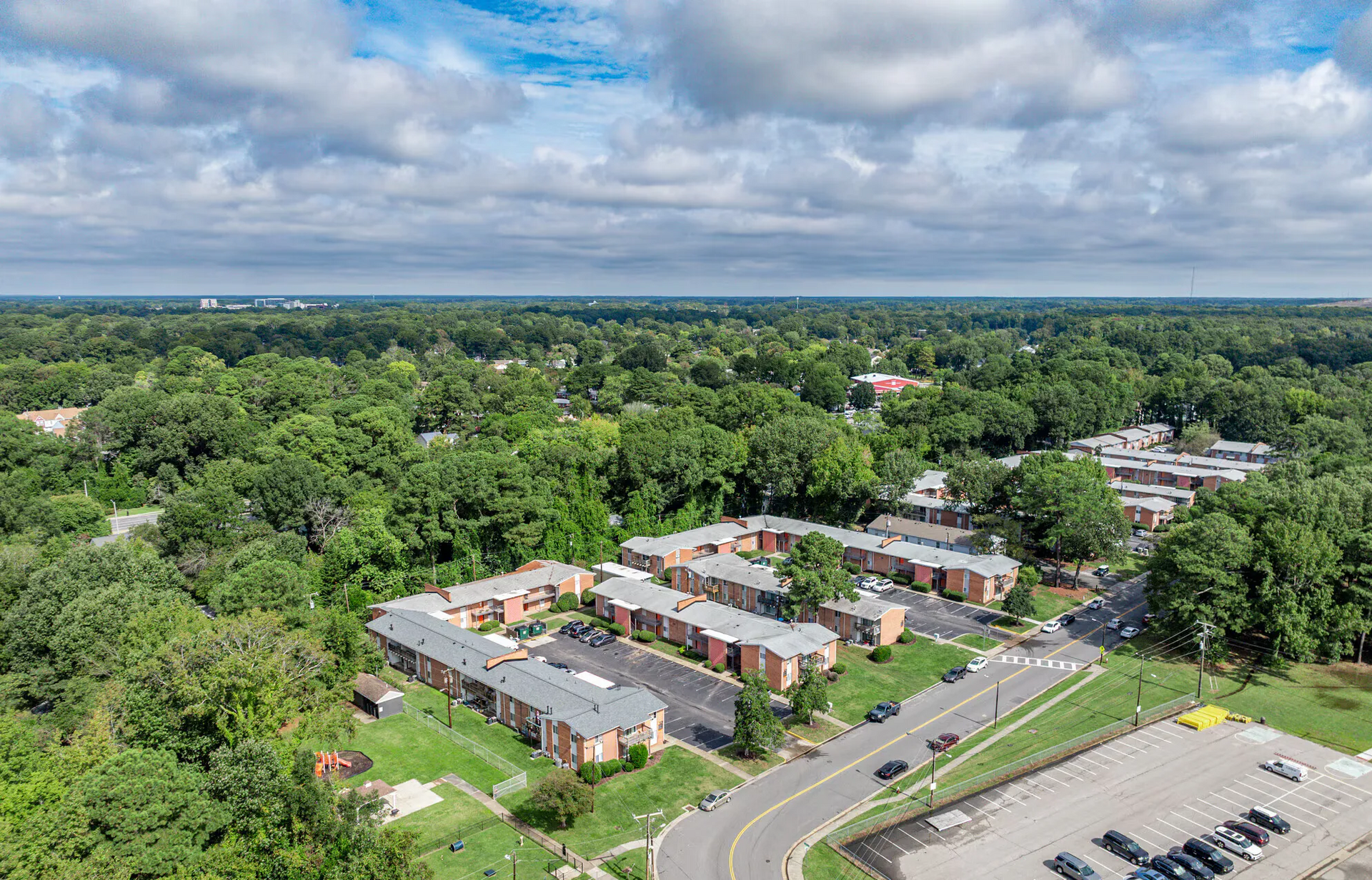 Aerial view of brick apartment buildings among trees with a parking lot. Welcome home to Harborstone Apartments, offering studio, 1, and 2-bedroom apartments for rent in New Port News, VA.