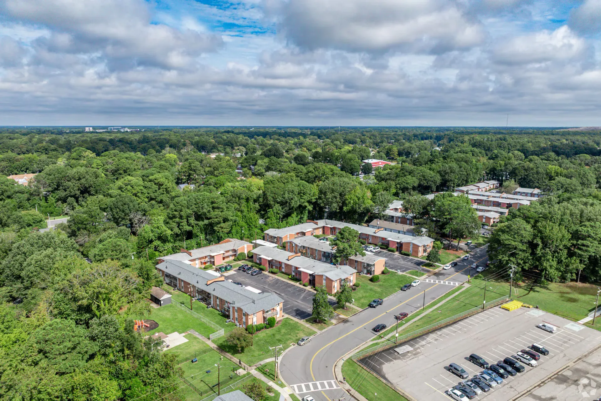 Aerial view of brick apartment buildings among trees with a parking lot. Welcome home to Harborstone Apartments, offering studio, 1, and 2-bedroom apartments for rent in New Port News, VA.