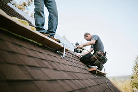 Two roofers kneel on a shingled roof, installing a gutter along the edge.
