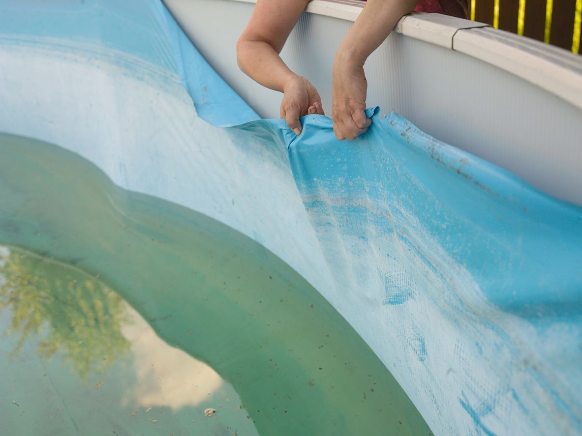 Hands pulling back a blue pool cover, revealing water in a backyard pool
